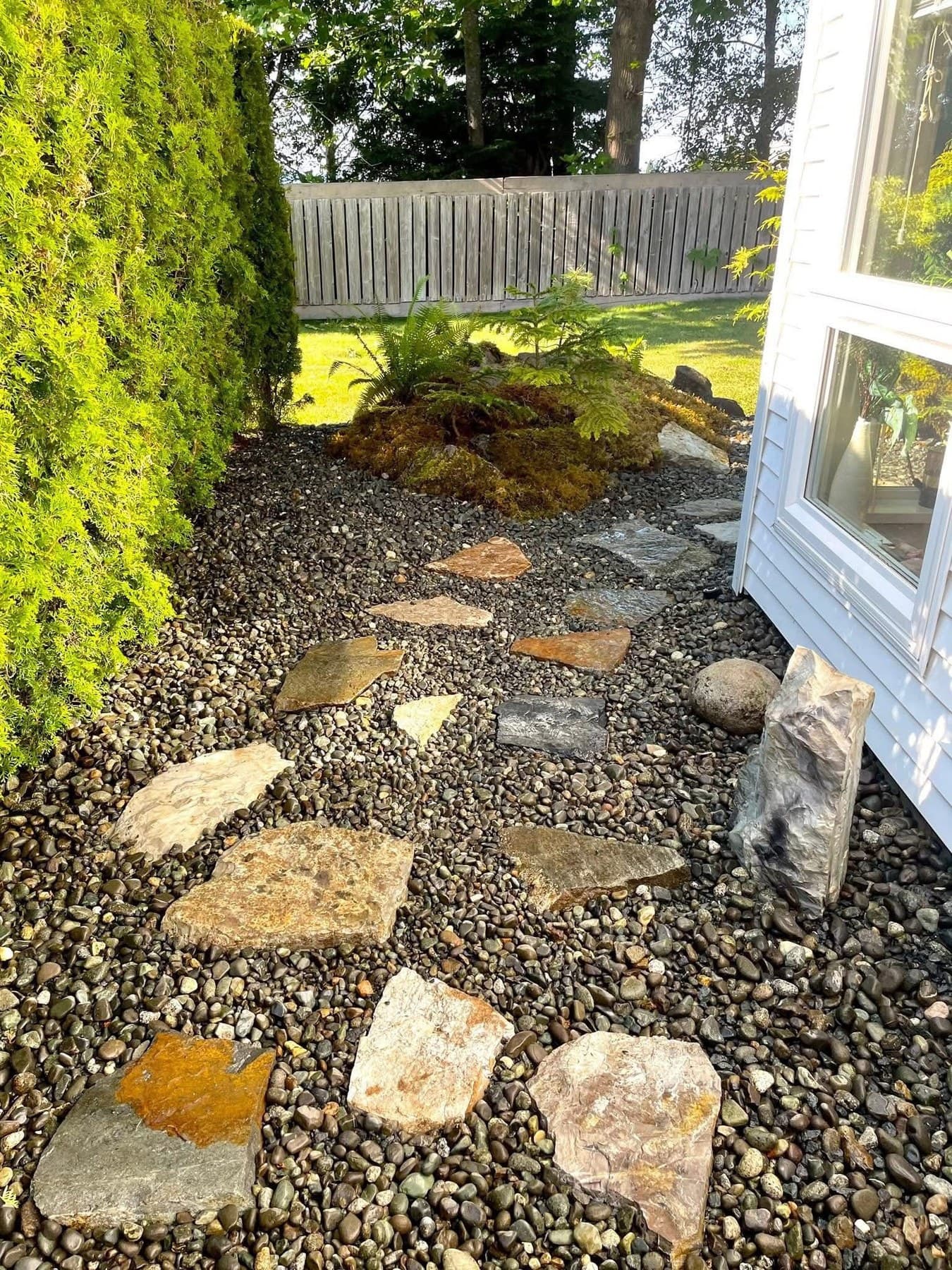 Stepping-stone flagstone path through river rock alongside a cedar hedge and moss-topped boulder