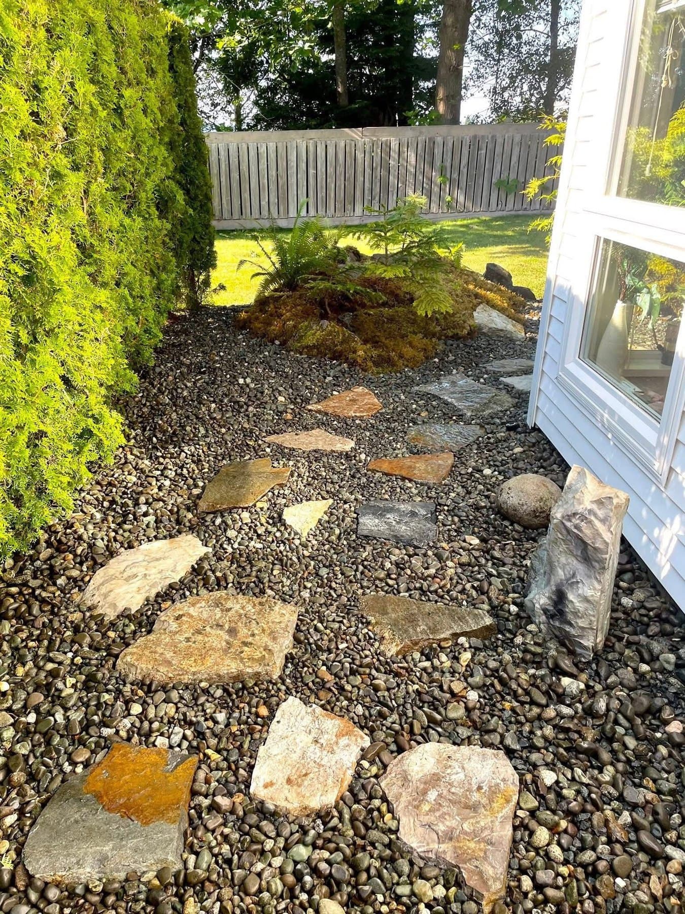 Flagstone stepping stones set into a river-rock pathway alongside a cedar hedge and moss-topped boulders