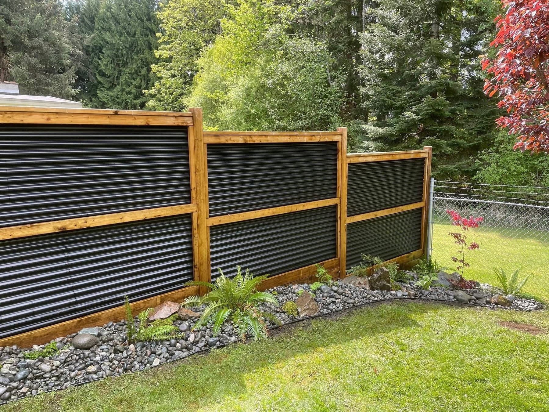 Modern wood and corrugated-metal privacy fence with a young Japanese maple, ferns, and river-rock border at the lawn edge